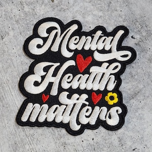 Puede incluir: Parche bordado blanco con el texto "Mental Health Matters" sobre un fondo negro. El texto está en una fuente cursiva. Hay tres corazones rojos y una flor amarilla bordada en el parche.
