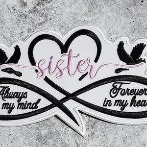 May include: Black and white embroidered patch with an infinity symbol and a heart. The heart has the word "sister" embroidered in pink. The infinity symbol has the words "Always on my mind" and "Forever in my heart" embroidered on each side.