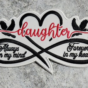 Pode incluir: Etiqueta branca bordada com um símbolo de infinito preto e um coração vermelho com a palavra "daughter" dentro. O símbolo de infinito tem as palavras "Always on my mind" e "Forever in my heart" de cada lado.