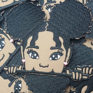 May include: A set of brown fabric patches featuring a cartoon girl with brown skin, black hair, and pink earrings. The girl is smiling and has her hands up, as if she is waving.