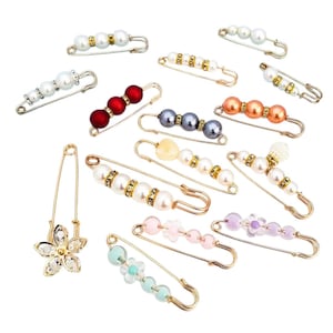 May include: A collection of gold-tone safety pins featuring various decorative elements including white, red, grey, orange, and pastel-colored beads, clear rhinestones, and a flower-shaped charm with clear crystals, arranged on a plain white background.