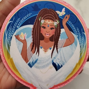 Puede incluir: Un parche redondo con borde de purpurina rosa sobre un fondo azul. En el centro, una ilustración de dibujos animados de una mujer negra con trenzas largas, vestida con un vestido blanco y una corona de flores. Tiene ojos azules y una tez marrón claro. La imagen está rodeada por un borde amarillo y naranja.