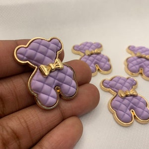 Op de afbeelding: Vijf paarse, gestikte knopen in de vorm van een teddybeer met een gouden rand en een gouden strik.