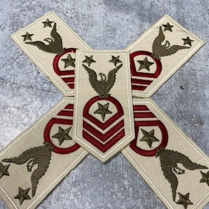Puede incluir: Cuatro parches de tela beige con estrellas, águilas y rayas bordadas en rojo, blanco y dorado. Los parches están dispuestos en forma de cruz.
