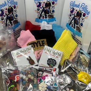 Könnte beinhalten: Eine Sock Star Bling Box gefüllt mit bunten Socken, Schmuck und Accessoires. Die Box ist mit einem Cartoon-Bild einer Frau verziert, die eine Krone trägt und eine Schere hält. Der Text "Sock Star Bling Box" ist auf der Box aufgedruckt. Die Box ist gefüllt mit einer Vielzahl von Artikeln, darunter Socken, Ohrringe, Halsketten und Anstecknadeln. Die Artikel sind alle in durchsichtigen Plastiktüten verpackt. Die Box ist ein tolles Geschenk für alle, die es lieben, Accessoires zu tragen.