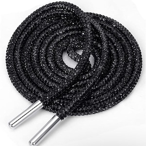 May include: Coiled black shoelaces, densely covered in small, glittering black crystals. The ends feature silver-coloured metal aglets. The crystals create a shimmering effect. These laces would add a touch of glamour to any pair of shoes, suitable for a special occasion.