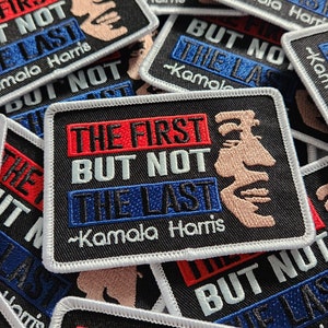 May include: A black fabric patch with white stitching around the edges. The patch features the text "The First But Not The Last - Kamala Harris" in red, white, and blue lettering. The text is in a bold, blocky font. A stylized image of Kamala Harris is on the right side of the patch.
