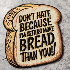 May include: Embroidered patch in the shape of a bread slice. The patch features the text "DON'T HATE BECAUSE I'M GETTING MORE BREAD THAN YOU!!" in black lettering on a cream-colored background, with a brown border.