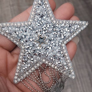May include: A silver star-shaped brooch, heavily embellished with sparkling crystals and rhinestones. A smaller black star is centered on the brooch. Silver beaded fringe hangs from the bottom of the star, adding movement and flair. The brooch is held in a hand.