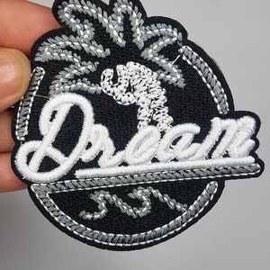Op de afbeelding: Zwarte geborduurde patch met een witte palmboom en het woord "Dream" in witte cursieve letters. De patch heeft een grijze gestikte rand.