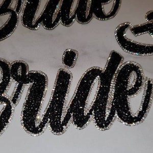 Op de afbeelding: Close-up van een zwarte strass "Bride" patch. De patch is gemaakt van zwarte strass steentjes op een zilveren ondergrond.