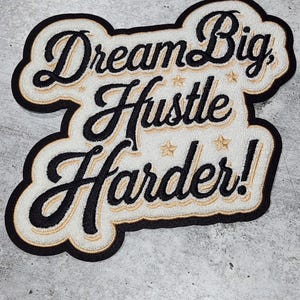 May include: White embroidered patch with black lettering that reads "Dream Big, Hustle Harder!" The patch has a black border and a white background.