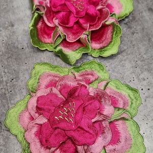 May include: Two pink and green embroidered fabric flowers. The flowers have a layered, ruffled design with a pink center and green outer edges.