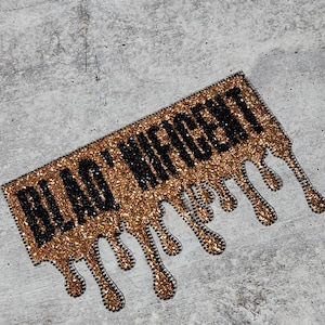 May include: A gold and black iron-on patch with the words "Black" and "Magic" in a dripping font. The patch is made of rhinestones and has a glittery finish.