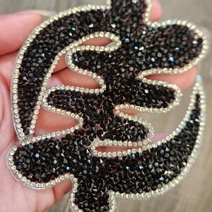 May include: A black and silver Adinkra symbol, made of rhinestones, representing Sankofa, meaning "return and get it".