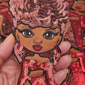 May include: A red and pink sequined iron-on patch of a cartoon woman with brown hair and a red dress.