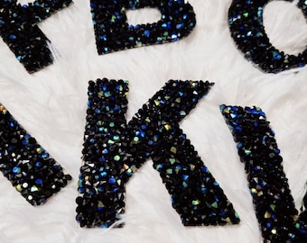 May include: Black letters of the alphabet covered in iridescent rhinestones. The letters are on a white fuzzy surface.