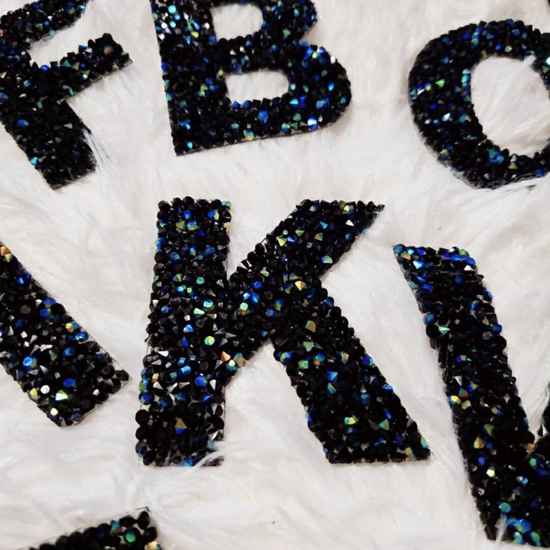 Peut inclure: Lettres noires de l'alphabet recouvertes de strass iris&eacute;s. Les lettres sont sur une surface blanche et duveteuse.