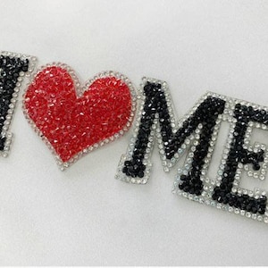 Könnte beinhalten: Dekorative Anordnung mit den Worten "I ❤️ ME" aus funkelnden schwarzen und roten Kristallen. Die Buchstaben und das Herz sind mit silberfarbenen Zierleisten umrandet, was ein glamouröses Design ergibt.