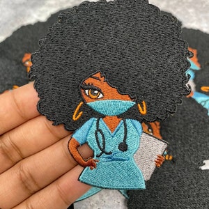 May include: Iron-on embroidered patch depicting a cartoon Black woman with an afro wearing a blue medical scrubs uniform, a face mask, and a stethoscope. She is holding a clipboard in her left hand.