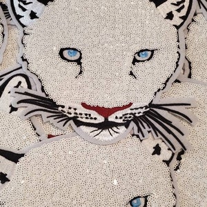 May include: A close-up of a white sequined fabric with a black and white embroidered design of a panther's face. The panther has blue eyes and a red nose.