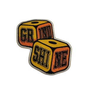 Pode incluir: Um emblema bordado com duas formas de dados em amarelo e laranja com bordas pretas. O dado superior exibe o texto GRIND e o dado inferior exibe o texto SHINE em letras pretas em negrito. O emblema tem uma borda costurada em cinza claro.