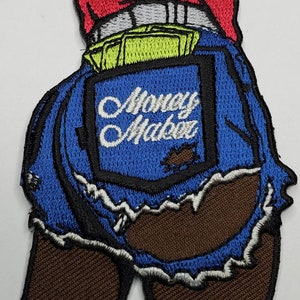 May include: An embroidered patch with a blue denim design. The patch features a back pocket with the text "Money Maker" and stacks of green bills. The denim is ripped and frayed around the edges.