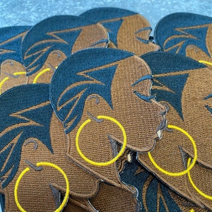 May include: A stack of brown fabric patches with embroidered images of a woman's face with yellow hoop earrings. The patches are layered on top of each other.