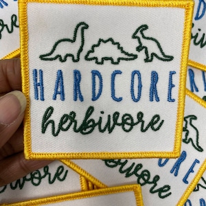 May include: A white embroidered patch with a yellow border. The patch features three green dinosaurs, the text "HARDCORE" in blue, and the text "herbivore" in green.