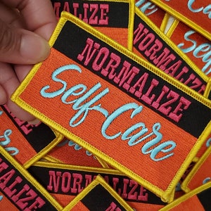 May include: A close-up of several embroidered patches with a black background and a red and yellow border. The patches say "Normalize Self-Care" in a white cursive font.