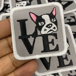 May include: A gray fabric patch with white stitching around the edges. The patch features a black and white illustration of a French bulldog with pink ears. The word "LOVE" is written in black letters below the dog.