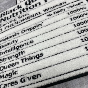 May include: White rectangular rug with black text that reads "Black Queen Nutrition Facts." The text includes phrases like "Melanin Drippin'," "Beauty," "Intelligence," and "Queen Things." The rug has a black border.