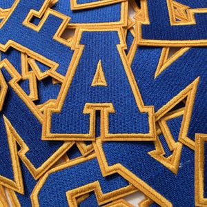 May include: A collection of embroidered letter patches in a varsity font style. Each patch features a royal blue center with a thick, stitched gold border. The letters are arranged in an overlapping pile, showcasing the textured thread detail.