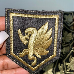 May include: A gold embroidered patch featuring a winged griffin design on a black felt background. The patch has a gold stitched border and is shaped like a shield.