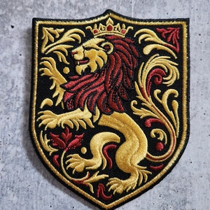 Può includere: Toppa ricamata raffigurante un leone d'oro rampante con criniera e corona rosse, su uno scudo nero. Il design include dettagli floreali dorati e rossi, incorniciati da un bordo nero. Adatta per personalizzare abbigliamento o accessori.