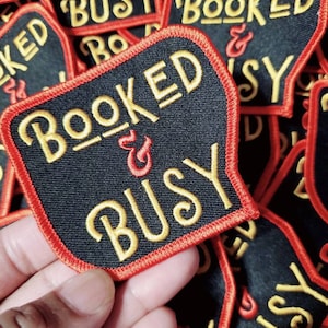 Pode incluir: Um remendo de tecido preto com costuras vermelhas ao redor das bordas. O remendo tem bordado dourado que diz "BOOKED & BUSY".
