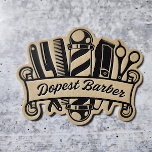 May include: A brown paper sticker with a black design of barber tools, including a razor, scissors, a comb, and a barber pole. The sticker says "Dopest Barber" in a black cursive font.