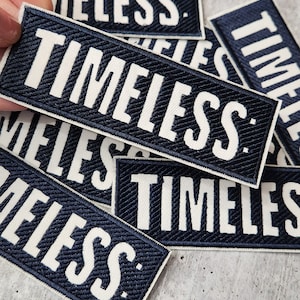 Può includere: Diversi patch in tessuto blu navy con testo bianco che recita "TIMELESS:".