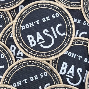 May include: Circular black patches with the words "DON'T BE SO BASIC" in white, surrounded by a tan border. The patches are embroidered and have a stitched edge. The patches are scattered across the image.