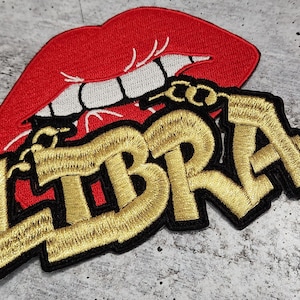 May include: Gold embroidered "LIBRA" text with a red, white, and black embroidered open mouth with teeth.