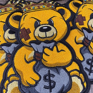 May include: A yellow embroidered teddy bear wearing a gold crown, holding a gray money bag with a dollar sign. The bear has a brown patch on its eye and a fierce expression.