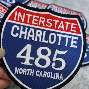 Puede incluir: Una señal de carretera interestatal azul y roja con texto blanco. La señal dice "Interstate Charlotte 485 North Carolina".