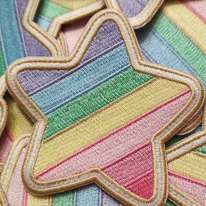 May include: Embroidered star patches in a rainbow of colors, including purple, blue, green, yellow, and pink. Each patch has a gold border and a white inner outline. The patches are scattered on a striped background.