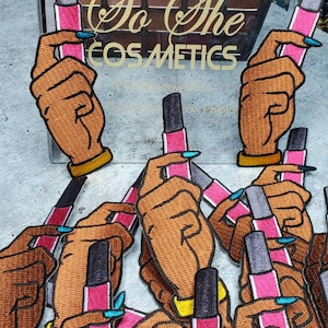 May include: A set of colourful embroidered patches featuring hands holding lipstick tubes. The patches are designed with a brown skin tone and feature the text "So She Cosmetics" in gold lettering.