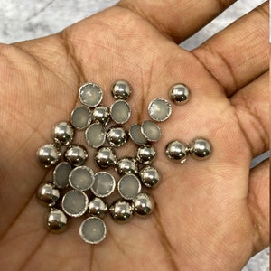 Könnte beinhalten: Eine Ansammlung kleiner, silberfarbener, halbrunder Perlen mit flacher, hellfarbiger Rückseite. Die Perlen sind über eine Hand verstreut und zeigen ihre reflektierende, metallische Oberfläche. Diese Perlen eignen sich ideal für Bastelarbeiten und Schmuckherstellung.