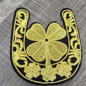May include: Gold embroidered horseshoe with a four-leaf clover in the center. The horseshoe is outlined in black and has a decorative design.