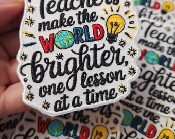 Op de afbeelding: Een witte geborduurde patch met de tekst "Teachers make the world brighter, one lesson at a time". De tekst is in zwart met een lampengrafiek in geel, rood, blauw en groen. De lamp heeft een wereldgrafiek erin.