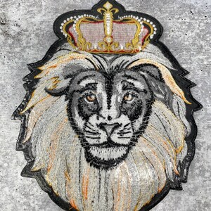 K&ouml;nnte beinhalten: Gestickter L&ouml;wenkopf-Patch mit einer goldenen Krone. Der L&ouml;we hat eine wei&szlig;e M&auml;hne und ein schwarzes Gesicht. Die Krone hat rote und goldene Akzente.
