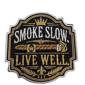 May include: A black embroidered patch with a white border, featuring the text "SMOKE SLOW." and "LIVE WELL." in gold and white. The design includes a central cigar illustration with a crown emblem, a top crown icon, and bottom laurel leaves.