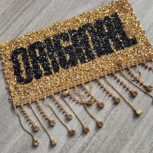 May include: A gold and black beaded patch with the word "remarkable" in black beads. The patch has a fringe of gold chains with beads.